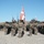 A group of military personnel in camouflage uniforms are posed in formation on a gravel terrain, flanked by armored military vehicles. Multiple national flags are visible, including what appears to be the Danish flag (red with white cross), a Georgian flag (white with red crosses), and a US flag. This appears to be a multinational military exercise or ceremonial formation.