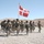 A formation of soldiers in full combat gear marches across a gravel terrain in an arid, desert-like environment. A flag bearing a red and white cross design — consistent with the flag of Oman — is carried at the front of the formation. The scene appears to be a military parade or exercise, likely in the Middle East based on the landscape.