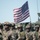 A group of heavily armed military personnel in full combat gear are assembled in formation with an American flag flying prominently in the background. The soldiers are equipped with tactical helmets, body armor, assault rifles, and face coverings, suggesting a formal military parade, exercise, or ceremonial event. A military vehicle is partially visible in the background to the right.