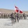 A multinational military parade or formation ceremony is taking place on an arid, gravelly terrain with hills in the background. Soldiers from multiple nations are marching in formation, with a prominent red-and-white flag (consistent with the Danish flag) being carried at the front. Multiple national flags are visible in the background, including what appear to be Georgian and American flags.