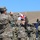 A military ceremony or parade is taking place outdoors in an arid, hilly landscape under a clear blue sky. Soldiers in camouflage uniforms are assembled in formation, with Georgian national flags visible. A conductor or commanding officer with raised arms appears to be directing a military band, with a bugler prominently visible in the foreground.
