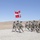 A column of military personnel in camouflage uniforms marches across an arid, desert-like terrain. A soldier at the front carries a flag featuring a white Nordic cross on a red background, consistent with the Danish flag design, along with an emblem. The formation appears to be a formal military march or parade exercise in an open, dry landscape.