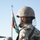 A soldier in camouflage uniform and helmet is photographed from behind/side profile, holding a rifle and wearing tactical gear including a backpack and knee pads. A shoulder patch displaying the Romanian tricolor flag (blue, yellow, red) and a unit insignia is clearly visible on the upper arm. A military flag on a pole is visible in the background against a clear blue sky.