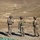 Three military personnel in camouflage uniforms are standing on an elevated rocky terrain, appearing to conduct a briefing or planning session. One soldier on the left is holding and reviewing documents, while the other two face him in discussion. The arid, semi-desert landscape with a winding dirt road is visible in the background below.