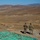 Three military personnel in camouflage uniforms stand on an elevated ridge overlooking a vast arid desert valley with rolling brown hills in the background. A large painted terrain map or sand table is spread on the ground in the foreground, suggesting a tactical planning or observation exercise. Several military vehicles are visible in the valley below, indicating an active field training exercise.