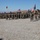 A military ceremony or parade is taking place in an arid, open terrain. A senior officer in a formal red ceremonial uniform with gold trim stands at attention in the foreground, facing a large formation of soldiers in combat fatigues. Multiple national flags are visible, including those of the United States and Georgia, along with armored military vehicles in the background, suggesting a multinational military exercise or joint training event.