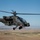 An AH-64 Apache attack helicopter is captured in flight over an arid, open landscape under a clear blue sky. The aircraft is painted in olive drab/dark green military camouflage and bears 'UNITED STATES' markings on the fuselage. At least one crew member is visible through the tandem cockpit canopy.