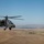 A military attack helicopter, identifiable as a Boeing AH-64 Apache, is captured in flight over an arid, semi-desert landscape with distant mountains and a flat valley visible in the background. The helicopter is painted in olive drab/army green camouflage and appears to be on a low-altitude flight mission. The image is taken from another airborne platform, suggesting a formation flight or aerial photography mission.