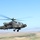 A military attack helicopter, identifiable as an AH-64 Apache, is captured in flight against a clear blue sky. The aircraft is photographed from another airborne platform, showing it in level flight over an arid, open landscape. The rotor blades show motion blur, indicating active flight.