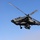 Two military attack helicopters are captured in flight against a clear blue sky. The foreground helicopter is prominently featured and appears to be an AH-64 Apache, showing visible weapons systems and sensor equipment. A second identical helicopter is visible in the lower right background, suggesting a formation flight.