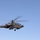 A military attack helicopter is captured in flight against a clear blue sky. The aircraft appears to be an AH-64 Apache, painted in olive drab/army green camouflage, with rotor blades in motion showing motion blur. A small red marking is visible on the fuselage near the center of the aircraft.