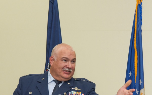 A senior U.S. Air Force officer in dress blue uniform is speaking or presenting at a formal military ceremony or event. He is gesturing with both hands in an expressive manner, standing between an American flag and a U.S. Air Force flag. Multiple service ribbons, medals, and pilot wings are visible on his uniform.