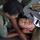 Two uniformed personnel are performing a first aid or medical training exercise on a person lying on the ground. One person is stabilizing the patient's head and jaw while another applies a cervical collar (neck brace). The uniforms display a patch that appears to read 'FIJI', suggesting this may be Fijian military or law enforcement training.
