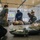 Military personnel are conducting a Tactical Combat Casualty Care (TCCC) training exercise in an indoor facility. A simulated casualty is lying on the floor while multiple trainees practice medical response techniques under instruction. A presentation screen in the background displays 'Three PHASES of TCCC' content.