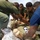 Several individuals in military/olive-green uniforms are performing what appears to be a medical training exercise on a person lying on a reflective emergency thermal blanket on the floor. The group is clustered around the casualty, applying hands-on treatment, suggesting a first aid or combat casualty care drill. One participant is wearing a blue civilian shirt, possibly indicating an instructor or observer role.