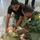 Two individuals are performing what appears to be a medical training exercise on a person lying on the floor wrapped in a mylar emergency blanket. The person in camouflage uniform with 'REMO' visible on the sleeve is attending to the casualty's neck/airway area, while a second person in a black shirt assists. The subject on the floor is wearing an olive military-style shirt with an 'Afghan' patch visible.