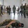 A military training session is taking place indoors, likely a Tactical Combat Casualty Care (TCCC) or combat first aid course, as indicated by the presentation screen showing 'Care Under Fire (CUF)', 'Tactical Field Care (TFC)', and 'Tactical Evacuation Care (TACEVAC)'. Personnel in mixed military uniforms — including US Army multicam and what appear to be Pacific Island nation military uniforms — are participating in a practical exercise. One soldier is demonstrating a low-crawl or casualty drag technique on the floor while others approach.