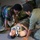 Two military personnel in uniform are providing first aid to a casualty lying on the floor. The patient is wearing a cervical collar (neck brace) and is partially covered with a mylar emergency thermal blanket. This appears to be a military medical training exercise or drill.