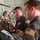 Three U.S. military personnel are gathered inside what appears to be a military transport aircraft, likely a C-130 or similar airlifter. Two individuals wearing olive drab flight suits and one soldier in Army combat uniform (ACU) with a helmet are reviewing documents and a tablet device. The setting suggests a mission briefing or pre-operation coordination taking place aboard the aircraft.