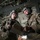 Two U.S. Air Force personnel in military flight gear are reviewing a document inside what appears to be a military transport aircraft. Both individuals are wearing flight helmets with communication headsets and OCP (Operational Camouflage Pattern) uniforms. Additional personnel are visible seated in the background of the aircraft's cargo bay.