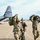 Three military personnel in OCP (Operational Camouflage Pattern) uniforms are walking across an airfield tarmac, carrying large green duffel bags. Behind them is a C-130 Hercules transport aircraft marked with 'ANG TEXAS' and tail number 85911, indicating it belongs to the Texas Air National Guard. The scene appears to depict soldiers offloading or departing after a flight.