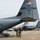 A Texas Air National Guard (ANG) C-130 Hercules transport aircraft, tail number 85911, is parked on a military flight line. Several soldiers in combat uniforms are loading or unloading gear from the aircraft's rear ramp. Additional aircraft, including what appear to be Polish and other allied military transports, are visible in the background.
