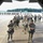 A large group of military paratroopers in full jump gear are staged on an airfield tarmac, viewed from inside the rear cargo bay of a military transport aircraft. The soldiers are wearing camouflage uniforms and carrying parachute packs, appearing to be preparing for a airborne operation or exercise. In the background, additional troops can be seen marching in formation across the tarmac, with other aircraft visible in the distance.