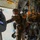 Military personnel are inside a transport aircraft, appearing to prepare for a parachute jump operation. Soldiers in camouflage uniforms and helmets are equipped with parachute packs and are connected to static lines. A jumpmaster or crew member is giving hand signals near the open aircraft door.
