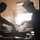 Two workers are preparing food in a commercial or institutional kitchen, with visible steam rising from the work surface. One worker wears a blue floral shirt and blue cap with gloves, while the other wears a black t-shirt with an American flag patch and a white cap with glasses. The dramatic backlighting from a bright window creates a high-contrast, atmospheric composition.