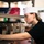 A young person wearing a white paper/cloth cap and black shirt is examining items on a commercial kitchen storage shelf. The shelf contains various large spice containers and food service supplies. The setting appears to be a restaurant or institutional kitchen storage area.