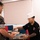 A military cook in black chef attire and white cap serves food to a Marine in digital camouflage uniform at what appears to be a military dining facility (chow hall). The cook, wearing glasses and displaying a tattooed forearm, is plating or serving food onto a tray. The setting is an institutional cafeteria-style environment with blue and red chairs visible in the background.