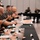 A group of military personnel in digital camouflage uniforms are seated along a long banquet table covered with a black tablecloth, applauding during what appears to be a formal military dining event or luncheon. The table is set with teal/turquoise trays, plates, bowls, and beverages, with partially eaten food visible. In the background, two individuals are seated at a separate table, likely serving as speakers or presiding officers for the event.