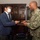 A U.S. Navy officer in NWU Type III camouflage uniform is exchanging business cards with a civilian man wearing a blue suit and white face mask. A second masked individual in a dark suit stands in the background. The setting appears to be a formal office with dark wood display cabinets containing awards and memorabilia.