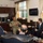 A formal meeting is taking place in what appears to be a military officer's office, with one person in U.S. Navy uniform speaking to four civilians in business suits. The setting suggests an official diplomatic or liaison meeting, indicated by the multiple international flags displayed on the cabinet in the background. The office is well-furnished with dark wood cabinetry, leather seating, and a view of trees through the windows.