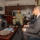 A U.S. Navy officer in digital camouflage uniform is meeting with three civilians in business attire in what appears to be a formal military office setting. The officer appears to be speaking and gesturing, suggesting an official briefing or diplomatic meeting. The setting includes international flags displayed on a credenza, indicating a command-level or liaison office.