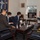 A formal meeting is taking place in what appears to be a military or government office, with four individuals seated in leather chairs around a coffee table. Three men in dark suits, two wearing face masks, are facing a fourth man in business casual attire who appears to be briefing or presenting information. A naval flag with an anchor and stars is displayed on the wall, and a computer monitor showing a presentation is visible in the background.