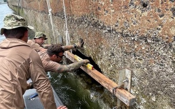 Three individuals in military-style tan uniforms and camouflage caps are working from a small aluminum boat alongside a large vessel or structure with a heavily fouled, barnacle-encrusted hull. They appear to be conducting a hull inspection or repair operation, using a wooden beam or plank positioned against an opening or damage point at the waterline. One person is reaching into or measuring a gap/hole in the hull while the others assist and stabilize.