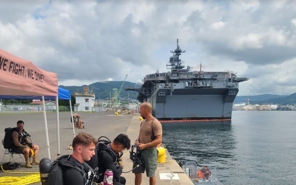 Military divers are preparing for a dive operation on a pier at what appears to be a naval base, with a large amphibious assault ship visible moored in the background. Several personnel in full scuba gear are gearing up near a tent bearing a partially visible motto reading 'BUILD, WE FIGHT, WE DIVE.' The setting appears to be a U.S. Navy installation, likely in Japan based on the surrounding terrain and infrastructure.