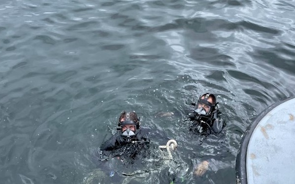 Two scuba divers are visible at the water surface next to what appears to be the hull of a vessel. Both divers are wearing full-face diving masks, black wetsuits, and diving equipment. One diver appears to be holding a rope or line near the boat's edge.