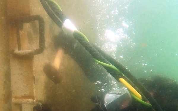 An underwater photograph showing a scuba diver working alongside a submerged concrete or stone structure with metal ladder rungs or attachment rings. The diver is equipped with full diving gear including a full-face mask branded 'OTS' and gloves labeled 'MOG'. The water has low visibility with a greenish tint, suggesting a harbor, pier, or industrial underwater environment.