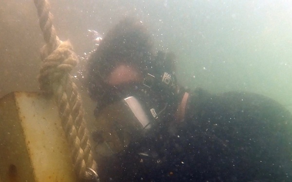 An underwater photograph showing a scuba diver in a black wetsuit working on or inspecting a submerged structure. The diver is in close proximity to what appears to be a metal frame or gate with a rope attached, along with cables. The water visibility is moderate with suspended particles visible throughout the image.