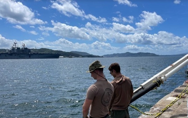 Two individuals stand on a concrete pier or dock, looking out toward open water where a large naval vessel is anchored in the background. A third person wearing a white hard hat, mask, and safety vest is visible to the right near what appears to be a large hose or cable running from the water onto the dock. The setting appears to be a military or industrial port facility with green hills visible in the distance.