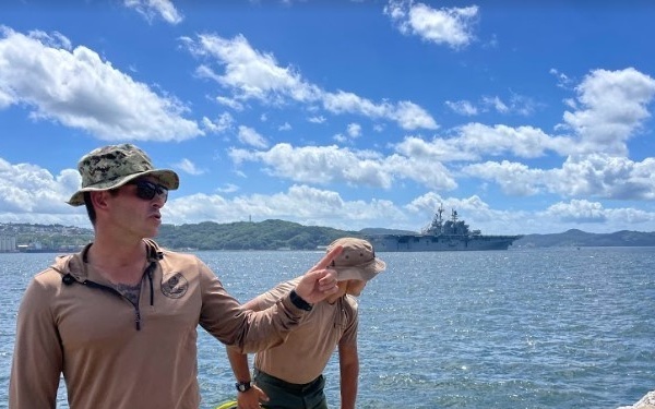 Two military personnel in tan/khaki uniforms and camouflage hats stand on a concrete pier or dock near a body of water. One individual appears to be pointing or gesturing while the other handles a yellow hose or cable. A large naval warship, likely an amphibious assault ship, is visible in the background on the water.