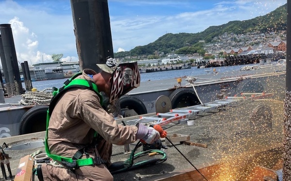 A worker in full personal protective equipment is performing grinding or cutting work on a metal structure at a waterfront facility, generating a shower of sparks. The setting appears to be a naval or industrial pier, with vessels and a hillside town visible in the background. The worker is kneeling on a concrete or coated surface and is secured with a green safety harness.
