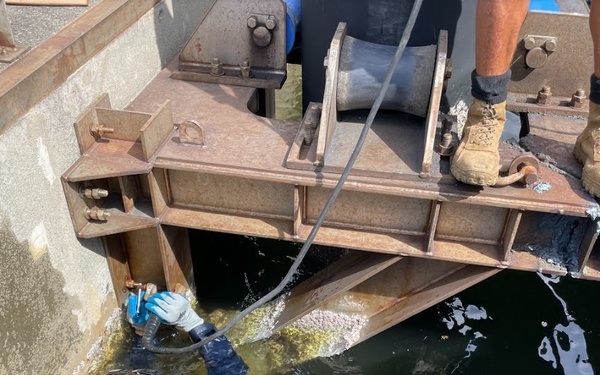 A diver in a wetsuit and snorkel mask is partially submerged in dark water, working on a large rusted steel structural assembly attached to what appears to be a dock or vessel hull. A second person, visible only from the knees down wearing tan boots and dark socks, is standing on the metal structure above. The diver appears to be performing underwater maintenance or inspection work, handling a hose or tool near the submerged metalwork.
