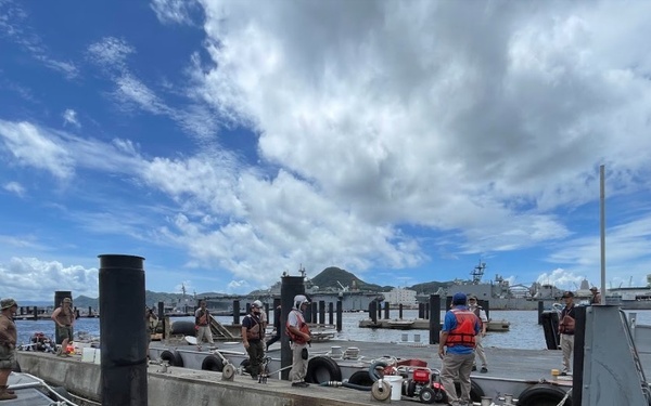 The image shows a military or naval working pier with multiple personnel conducting dockside operations. Several individuals wearing safety vests and work attire are visible on concrete floating dock sections, with larger naval vessels visible in the background harbor. The setting appears to be a tropical or subtropical naval installation based on the vegetation and cloud formations.
