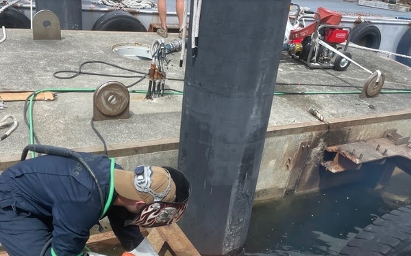 A worker in protective gear is performing welding or cutting operations on metal structural components aboard what appears to be a barge or work vessel. Sparks are visibly flying from the cutting/welding point on steel beams. The work is taking place in an open deck area near a water-filled compartment or well.