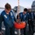 Three naval personnel in blue coveralls and inflatable life vests are working on a red marker buoy (numbered 402) on the deck of a military vessel. They appear to be conducting maintenance or inspection of the buoy's equipment, including electronic components mounted on top. The scene takes place at dusk or dawn, with open ocean visible in the background alongside naval superstructure.
