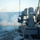 A close-in weapon system (CIWS), identifiable as a Phalanx Block 1B, is actively firing from the deck of a US Navy warship at sea. A visible plume of white gun smoke extends to the left of the barrel, indicating the weapon has just discharged. The ship appears to be underway on calm, open ocean under clear skies.
