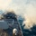 An aerial view of a naval warship, likely a U.S. Navy guided-missile destroyer, firing its forward deck gun during a live-fire exercise at sea. A large plume of white smoke is visible emanating from the gun muzzle toward the starboard side, indicating a recent or ongoing discharge. Several crew members are visible on the forward deck near equipment and what appears to be a helicopter landing pad.